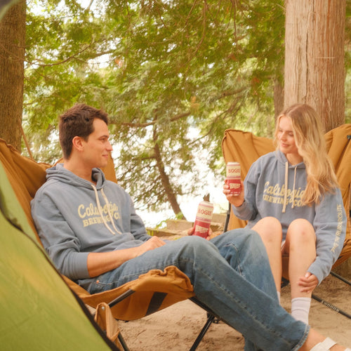 A man and a woman sitting in tan camping chairs inside a tent, wearing matching light blue hoodies with 'Calabogie Brewing Co.' printed in cream across the chest, surrounded by trees outdoors.