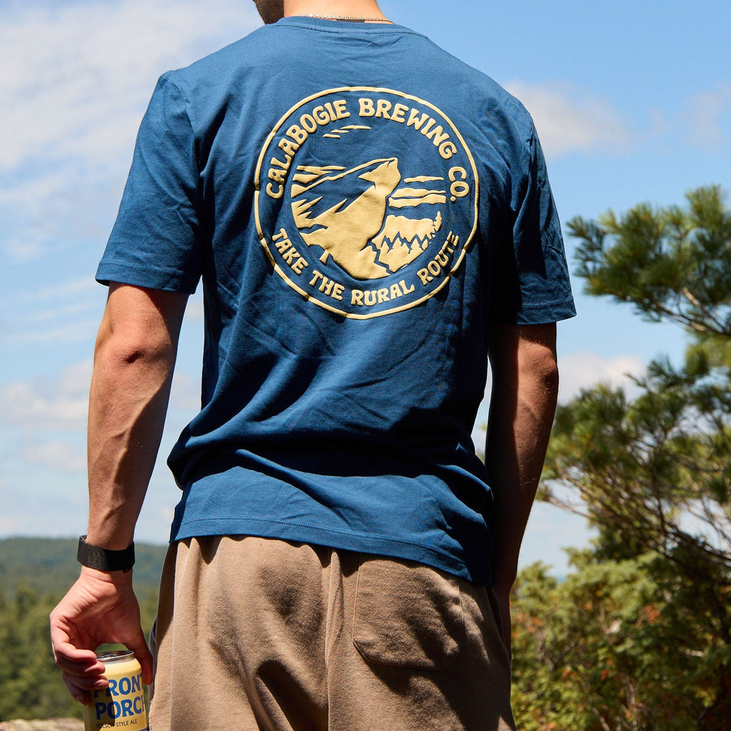 A man viewed from behind wearing a blue crewneck t-shirt with a large circular yellow graphic on the back that reads 'Calabogie Brewing Co. Take the Rural Route' with a duck illustration.