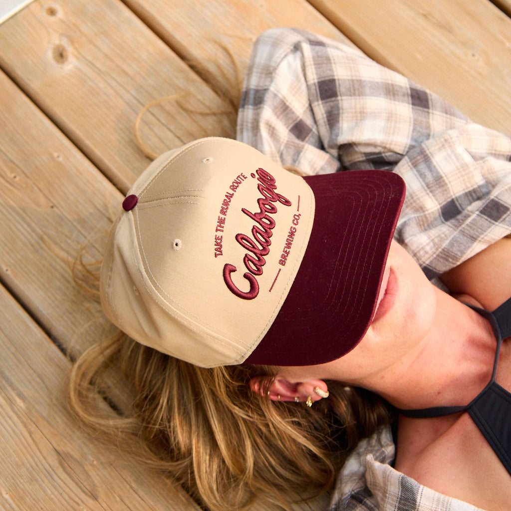 A woman lying on a wooden deck wearing a beige and burgundy baseball cap with 'Calabogie' embroidered on the front.